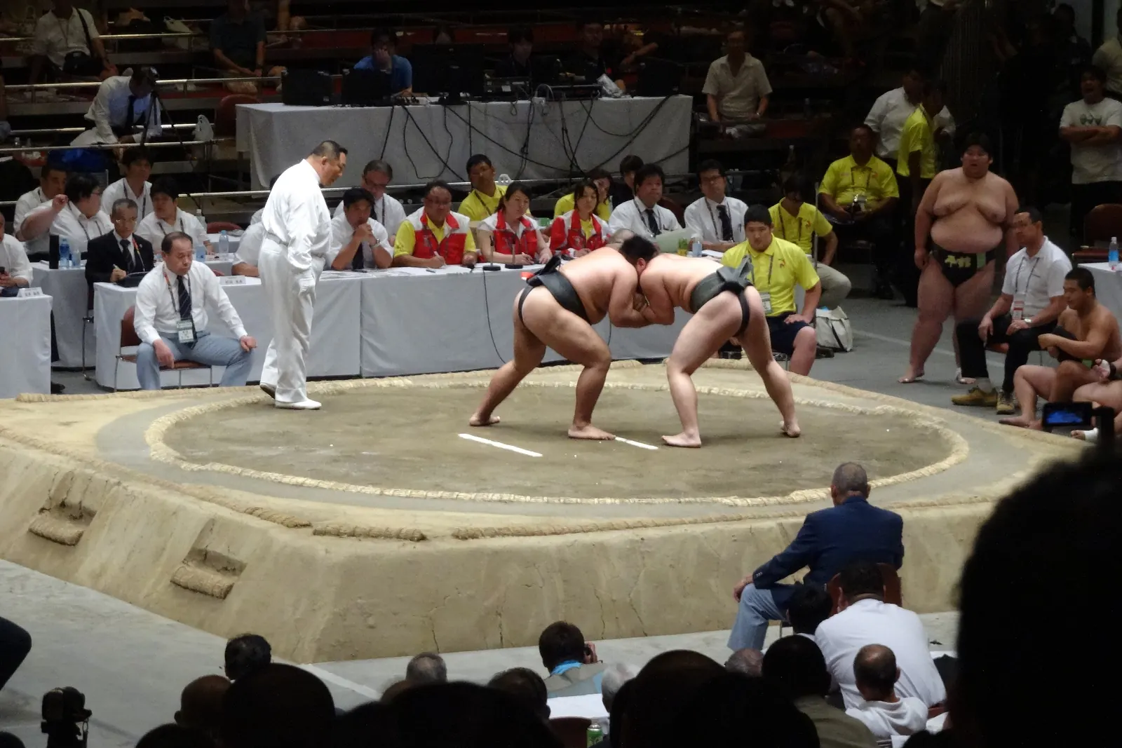 Two amateur sumo wrestlers lock together in a clay ring while a referee in white crouches beside them and people sit at a long table behind them