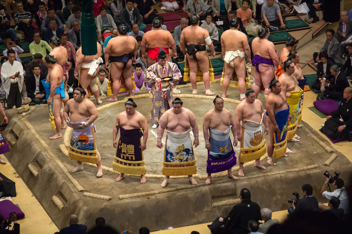 Rows of sumo wrestlers in decorated ceremonial aprons stand in a ring around the edge of a clay ring, photographed from above