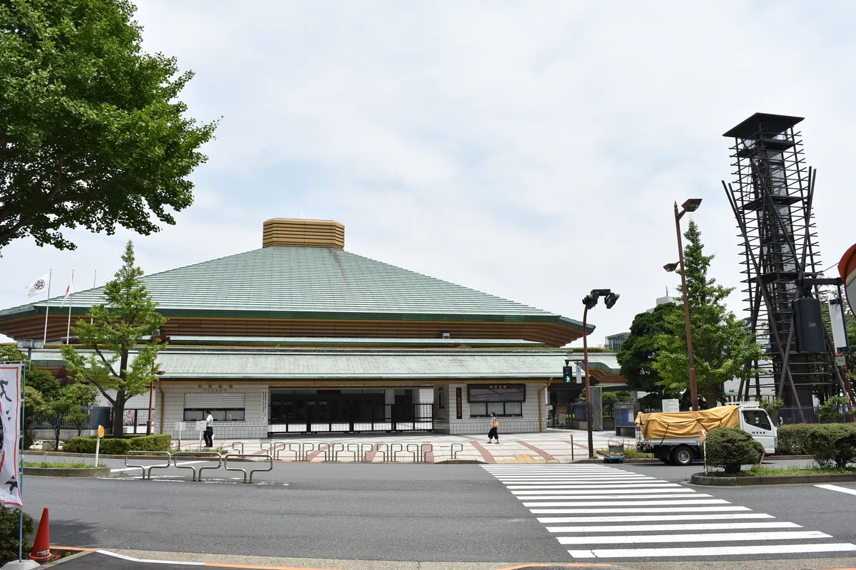 The low green roof of the Ryōgoku Kokugikan sumo hall stands behind trees and a pedestrian crossing under a cloudy sky