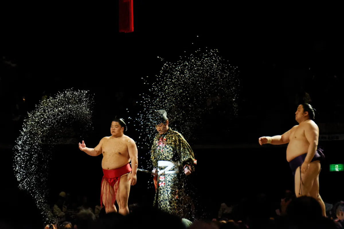 Two sumo wrestlers and a referee in a gold robe stand in darkness as arcs of thrown salt glitter in the air around them