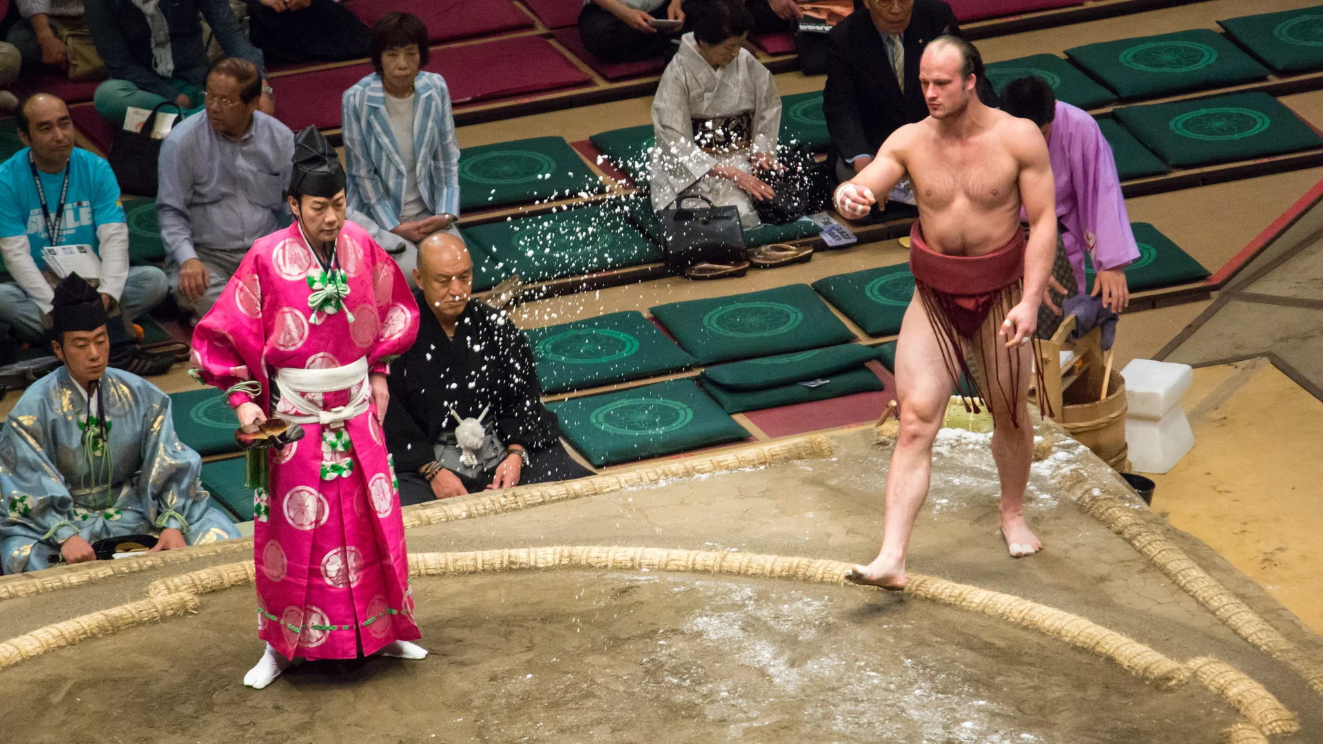 A sumo wrestler in a red belt throws salt into a clay ring at a Tokyo tournament while a referee in pink robes stands beside him
