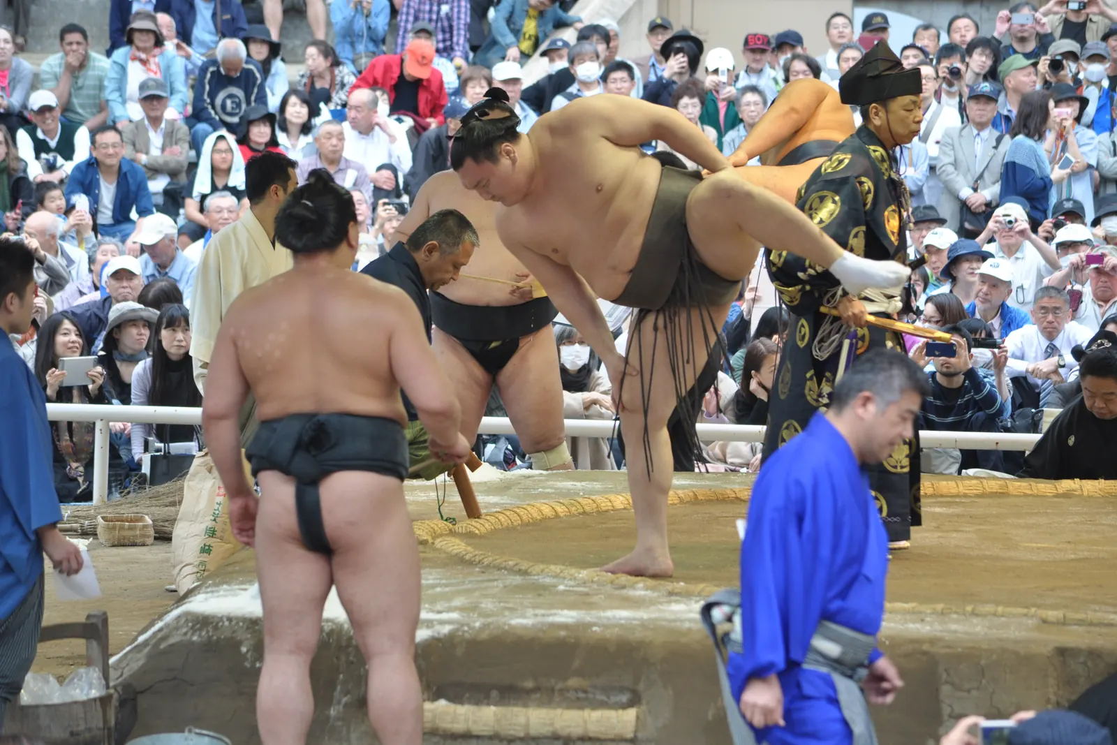 A sumo wrestler lifts one leg high beside the ring while other wrestlers and a referee stand around him and a crowd looks on